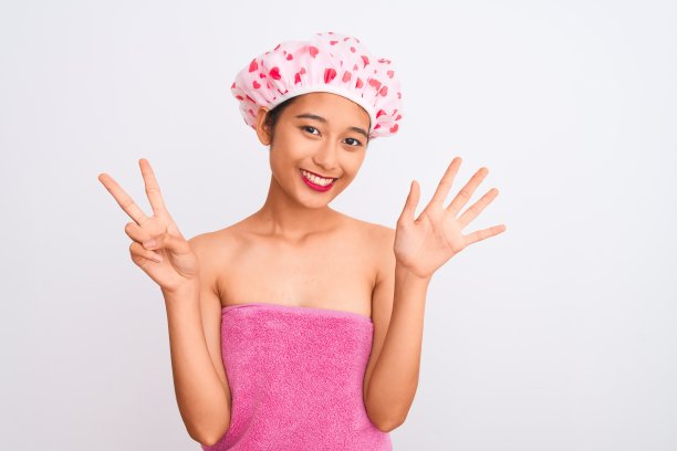 年轻的中国女子穿着浴巾和浴帽，在孤立的白色背景下，用七号手指指着前方，自信而快乐地微笑着。图片下载