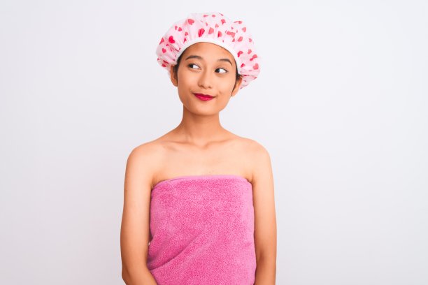 年轻的中国妇女穿着浴巾和帽子浴在孤立的白色背景微笑看着一边和凝视远方思考。图片下载