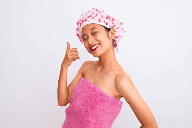 年轻的中国女子穿着浴巾和帽子沐浴在孤立的白色背景，微笑着做电话手势的手和手指，就像在打电话。沟通的概念。图片下载