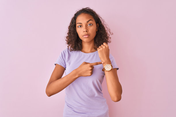 年轻的巴西女子穿着t恤站在孤立的粉色背景下，匆忙地指着手表时间，不耐烦地看着镜头，用轻松的表情图片下载