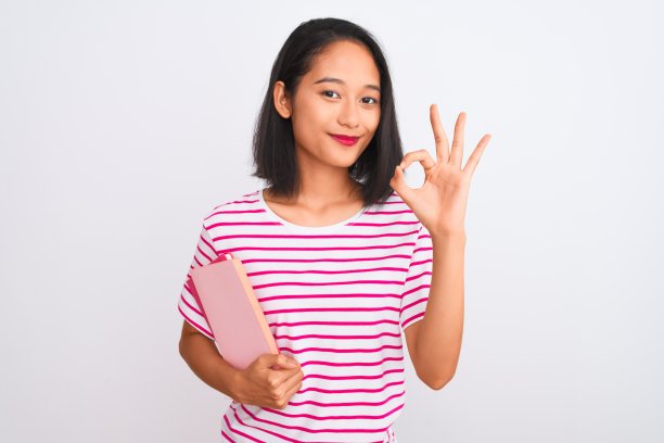年轻美丽的中国女人拿着书站在孤立的白色背景做ok手势的手指，优秀的象征图片下载