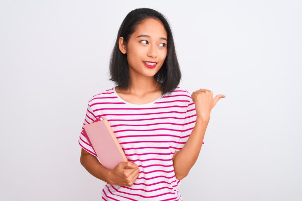 年轻美丽的中国女人捧着书站在孤立的白色背景指向和显示拇指向上的一边与幸福的脸微笑图片下载