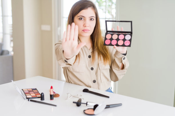 美丽的年轻女子使用化妆化妆品应用颜色从调色板与开放的手做停止标志与严肃和自信的表情，防御手势图片下载