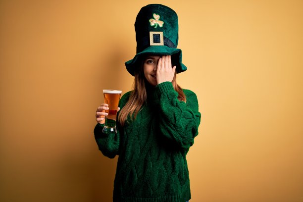 年轻美丽的女人戴着绿帽子喝一杯啤酒在圣帕特里克节覆盖一只眼睛与手，自信的微笑在脸上和惊讶的情绪。图片下载