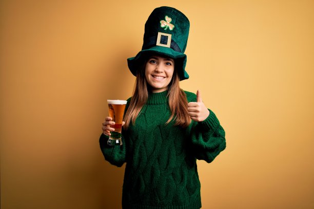 年轻美丽的女人戴着绿帽子喝啤酒在圣帕特里克节做快乐的拇指手势。赞许的表情看着镜头，表示成功。图片下载