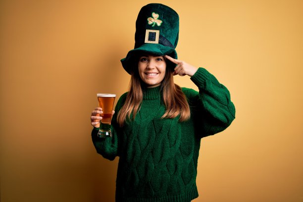 年轻美丽的女人戴着绿帽子喝一杯啤酒在圣帕特里克节用一个手指指着头微笑，伟大的主意或想法，良好的记忆图片下载