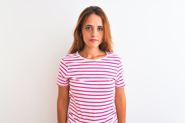 年轻的红发女子穿着条纹休闲t恤站在白色孤立的背景轻松与严肃的表情在脸上。简单自然的看相机。图片下载