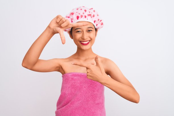 年轻的中国女人穿着浴巾和帽子浴在孤立的白色背景微笑做框架与手和手指与快乐的脸。创意和摄影概念。图片下载