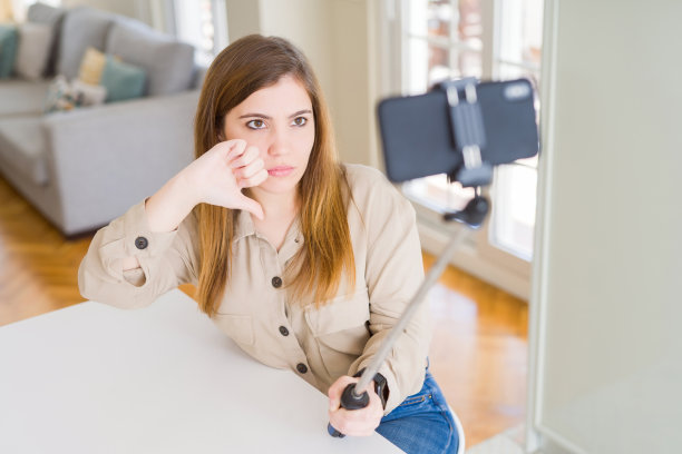 美丽的年轻女子用自拍杆拍照，脸上带着愤怒，消极的信号表示不喜欢，拇指向下，拒绝的概念图片下载