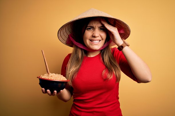 年轻女子戴着亚洲帽，吃着中国面条在黄色孤立的背景强调与手在头上，震惊与羞愧和惊讶的脸，愤怒和沮丧。为错误而恐惧和不安。图片下载