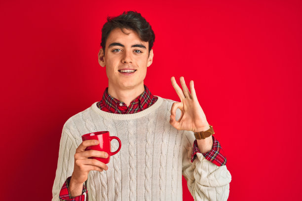 十几岁的男孩穿着冬季毛衣喝一杯咖啡在孤立的红色背景做ok手势与手指，优秀的象征图片下载