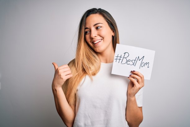 年轻美丽的女人拿着纸与最好的妈妈消息庆祝母亲节指向和显示与拇指向上的一边与快乐的脸微笑图片下载