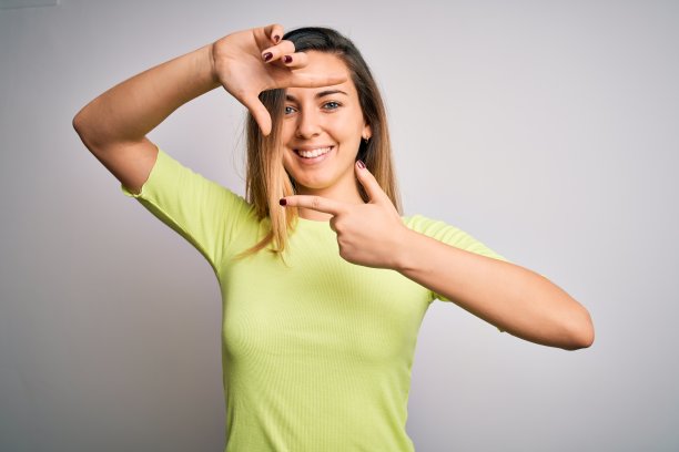 美丽的金发女人与蓝色眼睛穿着绿色休闲t恤在白色的背景微笑，使框架与手和手指与幸福的脸。创意和摄影概念。图片下载