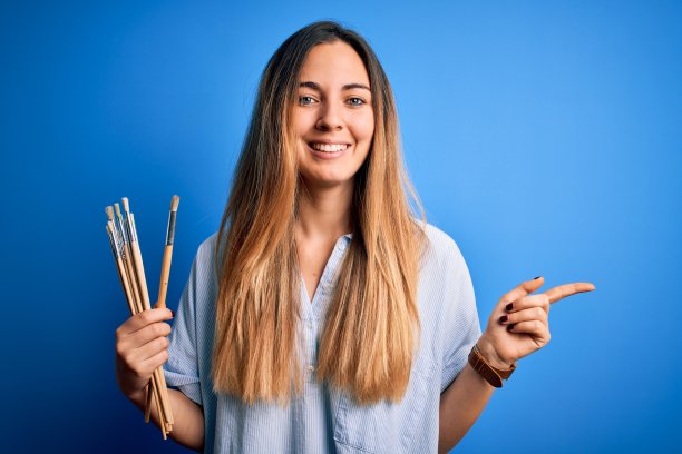年轻美丽的金发艺术家女人与蓝色眼睛绘画用画笔非常幸福的指向与手和手指的一边图片下载