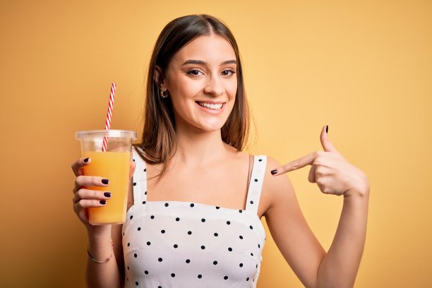 年轻美丽的黑发女人喝健康的橙汁黄色背景与惊讶的脸指向自己图片下载