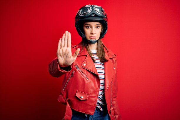 年轻美丽的黑发女摩托车手戴着摩托车头盔和红色夹克做停止唱歌与手掌。警告表情与否定和严肃的手势在脸上。图片下载