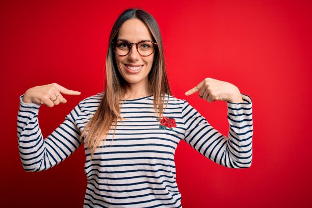 年轻美丽的金发女人与蓝色的眼睛戴眼镜站在红色的背景看起来自信的微笑在脸上，用手指指着自己骄傲和幸福。图片下载