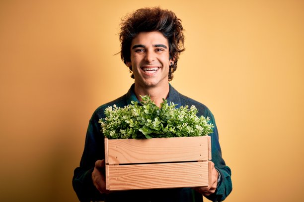年轻英俊的男子抱着盒子与花园植物在孤立的黄色背景与快乐的脸站着，微笑着自信的微笑显示牙齿图片下载