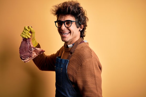 年轻英俊的屠夫男人抱着满足牛排站在孤立的黄色背景看向一边，放松的侧面姿势与自然的脸和自信的微笑。图片下载