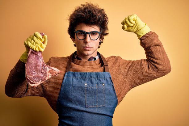 年轻英俊的屠夫捧着牛排站在孤立的黄色背景下强壮的人显示手臂肌肉，自信和自豪的力量图片下载
