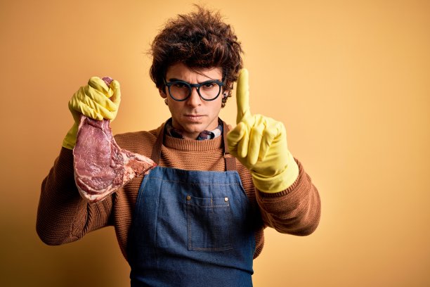 年轻英俊的屠夫手捧着肉排站在孤立的黄色背景下，愤怒地竖起手指，没有任何手势图片下载