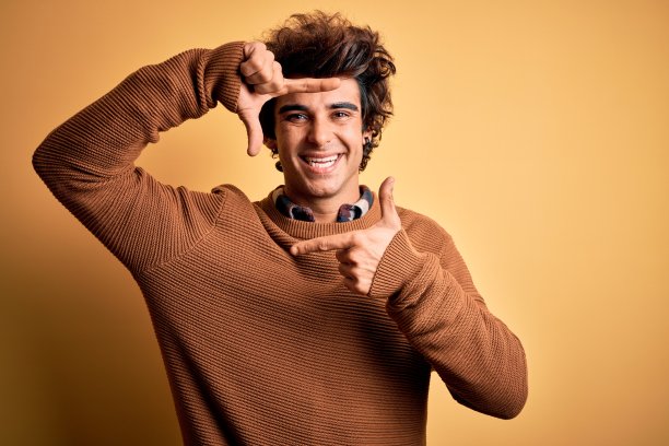 年轻英俊的男子穿着休闲衬衫和毛衣在孤立的黄色背景微笑做框架与手和手指与幸福的脸。创意和摄影概念。图片下载