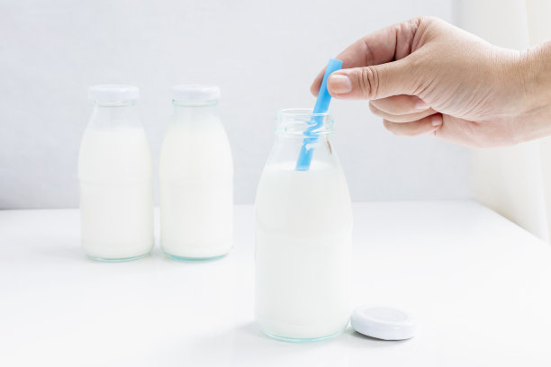 女人一手拿着蓝色的吸管，玻璃瓶里装着牛奶，桌上白色的背景为食物和健康的概念。与复制空间的文本。图片下载