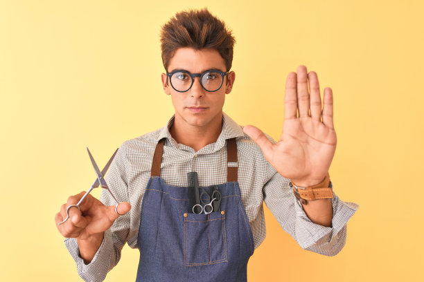 英俊的理发师穿着围裙拿着剪刀在孤立的黄色背景与开放的手做停止标志与严肃和自信的表情，防御手势图片下载