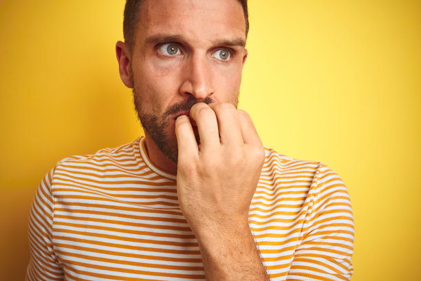 年轻英俊的男子穿着休闲t恤在黄色孤立的背景看起来紧张和紧张的手在嘴咬指甲的特写。焦虑的问题。图片下载
