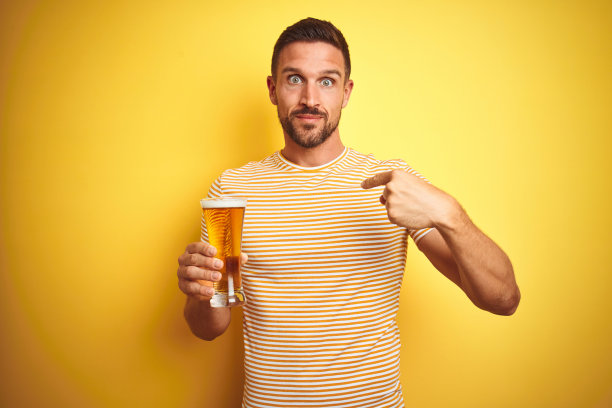 年轻英俊的男子喝一品脱啤酒在孤立的黄色背景与惊讶的脸指向自己图片下载