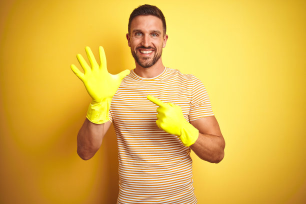 年轻英俊的男子戴着清洁手套为家务孤立的黄色背景非常幸福的指指点点图片下载
