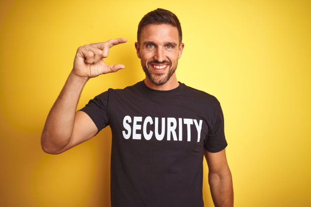 年轻的保安人员穿着安全制服在黄色孤立的背景微笑和自信的手势与手做小尺寸的标志，手指看着和相机。测量的概念。图片下载