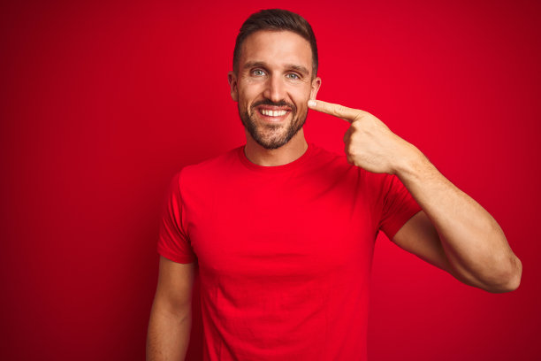 年轻帅气的男子穿着休闲t恤越过红色孤立的背景，用手手指着脸和鼻子，笑嘻嘻的。美丽的概念图片下载