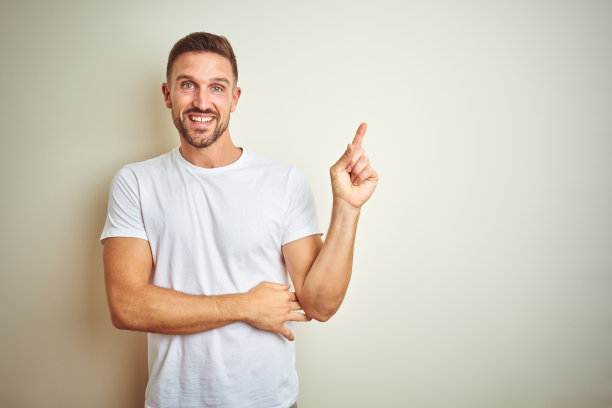 年轻帅气的男子穿着休闲白色t恤越过孤立的背景，脸上带着大大的微笑，用手指指着一边看着镜头。图片下载