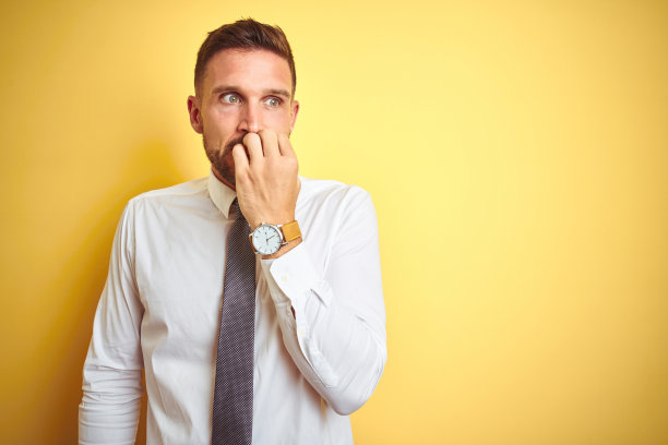 年轻英俊的商人穿着优雅的白色衬衫在黄色孤立的背景看起来紧张和紧张的手在嘴咬指甲。焦虑的问题。图片下载