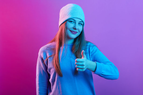 一位喜悦迷人的年轻女性戴着帽子和运动衫的照片，竖起大拇指，举起手，做出手势，直视镜头，面部表情愉快，表示赞同。图片下载