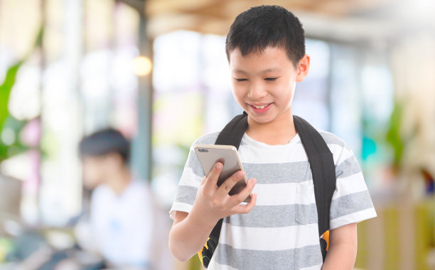 快乐的亚洲男孩背着书包在模糊的教室里使用智能手机。人们使用科技的概念图片下载