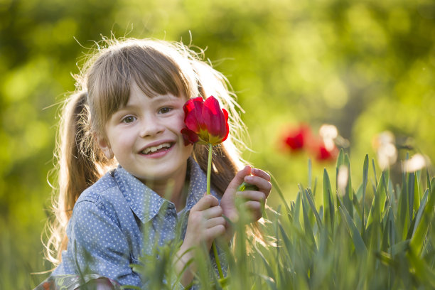 可爱的漂亮微笑的孩子女孩与灰色的眼睛和长头发与明亮的红色郁金香花在模糊阳光下的绿色散焦背景。热爱自然的理念。图片下载