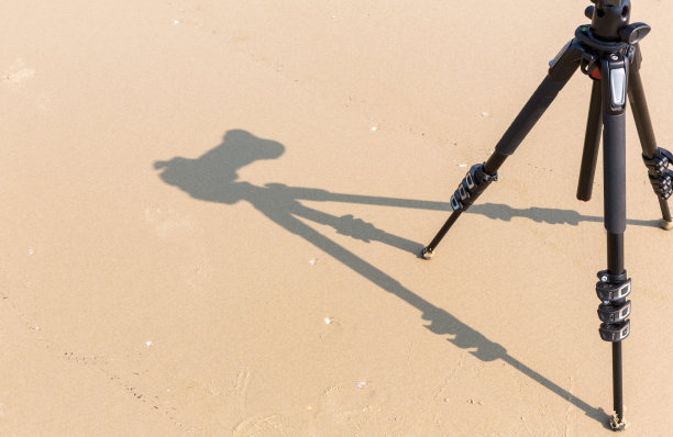 三脚架上相机的影子。以沙滩为背景的拍摄过程。从上面看到一个三脚架阴影的相机在沙滩上的影子。图片下载