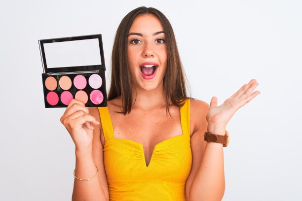 年轻美丽的女孩拿着化妆品站在孤立的白色背景非常高兴和兴奋，胜利者的表情庆祝胜利尖叫着大大的微笑和举起手图片下载