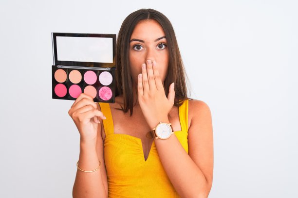 年轻美丽的女孩捧着妆站在孤立的白色背景上，用手捂住嘴，为错误而震惊，表情恐惧，在沉默中害怕，秘密的概念图片下载