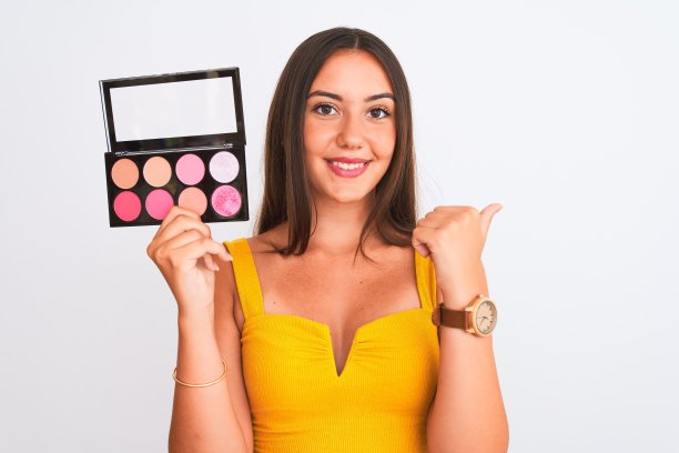 年轻美丽的女孩拿着化妆品站在孤立的白色背景指向和显示拇指向上的一边与幸福的脸微笑图片下载