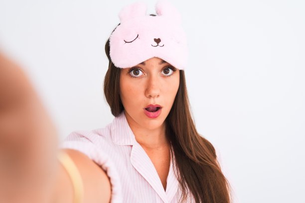 女孩穿着睡衣，戴着睡眠面罩，在孤立的白色背景下，用相机自拍，惊恐地看着惊讶的脸，害怕和兴奋的恐惧表情图片下载