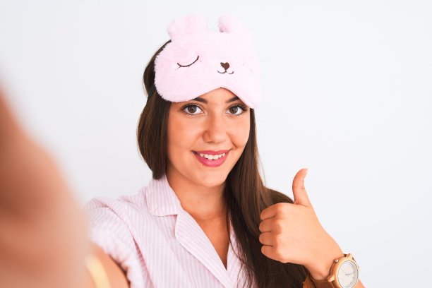 女孩穿着睡衣，戴着睡眠面罩，在孤立的白色背景下，用相机自拍，笑容满面，做着“ok”的手势，竖起大拇指，很棒的手势图片下载