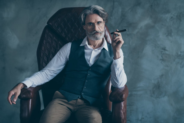 富人的肖像富有的成功优雅的惊人的老人黑手党领导人坐扶手椅持有雪茄看起来华丽的穿着背心衬衫棕色裤子裤子孤立的灰色背景图片下载