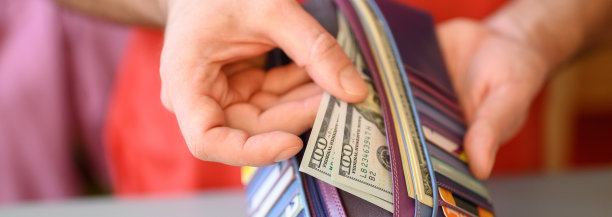 男人的手拿着100美元的钞票和一个钱包放在灰色的桌子上。横幅图片下载