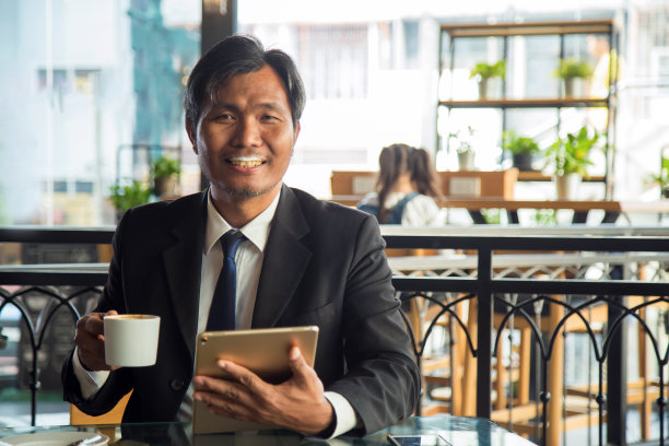 近距离的中年商人在正式的西装拿着一个咖啡杯看着镜头与牛奶泡沫在他的嘴在咖啡店。图片下载