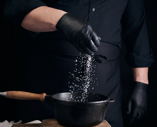 男厨师穿着黑色制服，戴着乳胶手套，在黑色铸铁平底锅里腌制食物图片下载