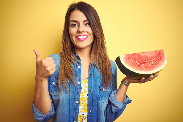 年轻漂亮的女人吃新鲜健康的西瓜片在黄色的背景高兴地与大微笑做ok的手势，拇指与手指向上，优秀的手势图片下载