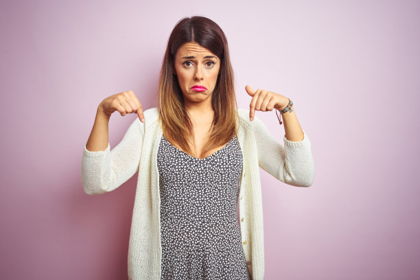 年轻美丽的女子站在粉色孤立的背景，向下看着悲伤和不安，用手指指示方向，不开心和沮丧。图片下载
